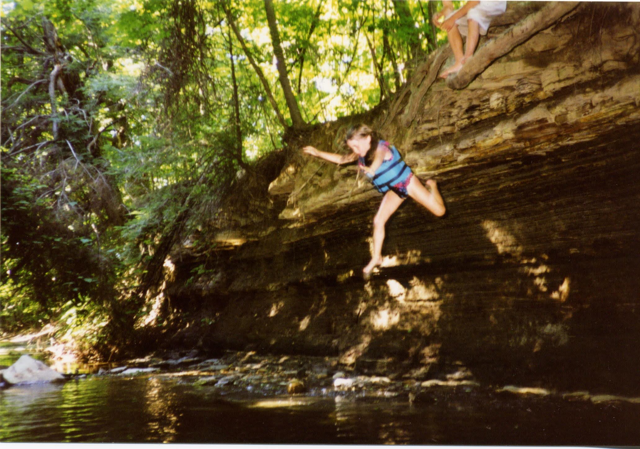 Christina in a blue life vest leaping off a creek-side cliff into the water below, mid-jump, around 1996