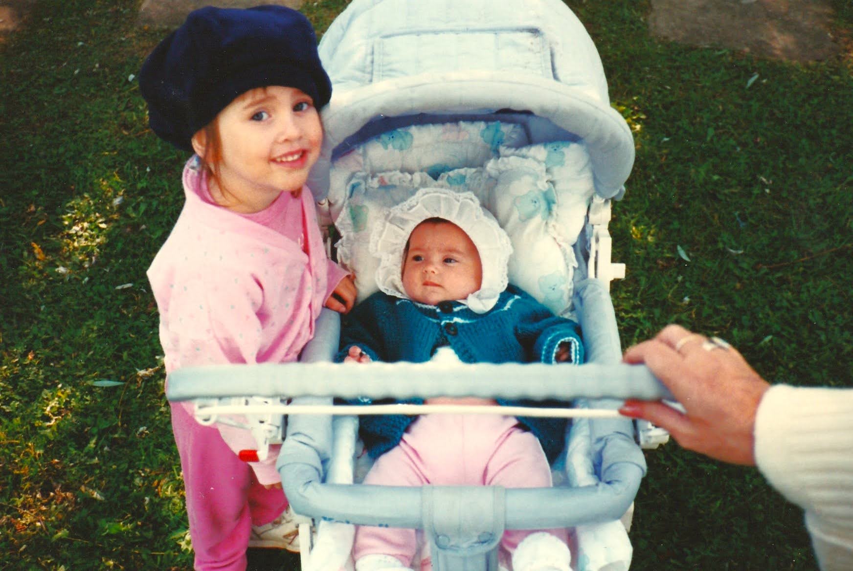 Toddler Christina in a navy beret and pink coat smiling next to baby Kim in a stroller with a white bonnet and teal knit sweater, 1994