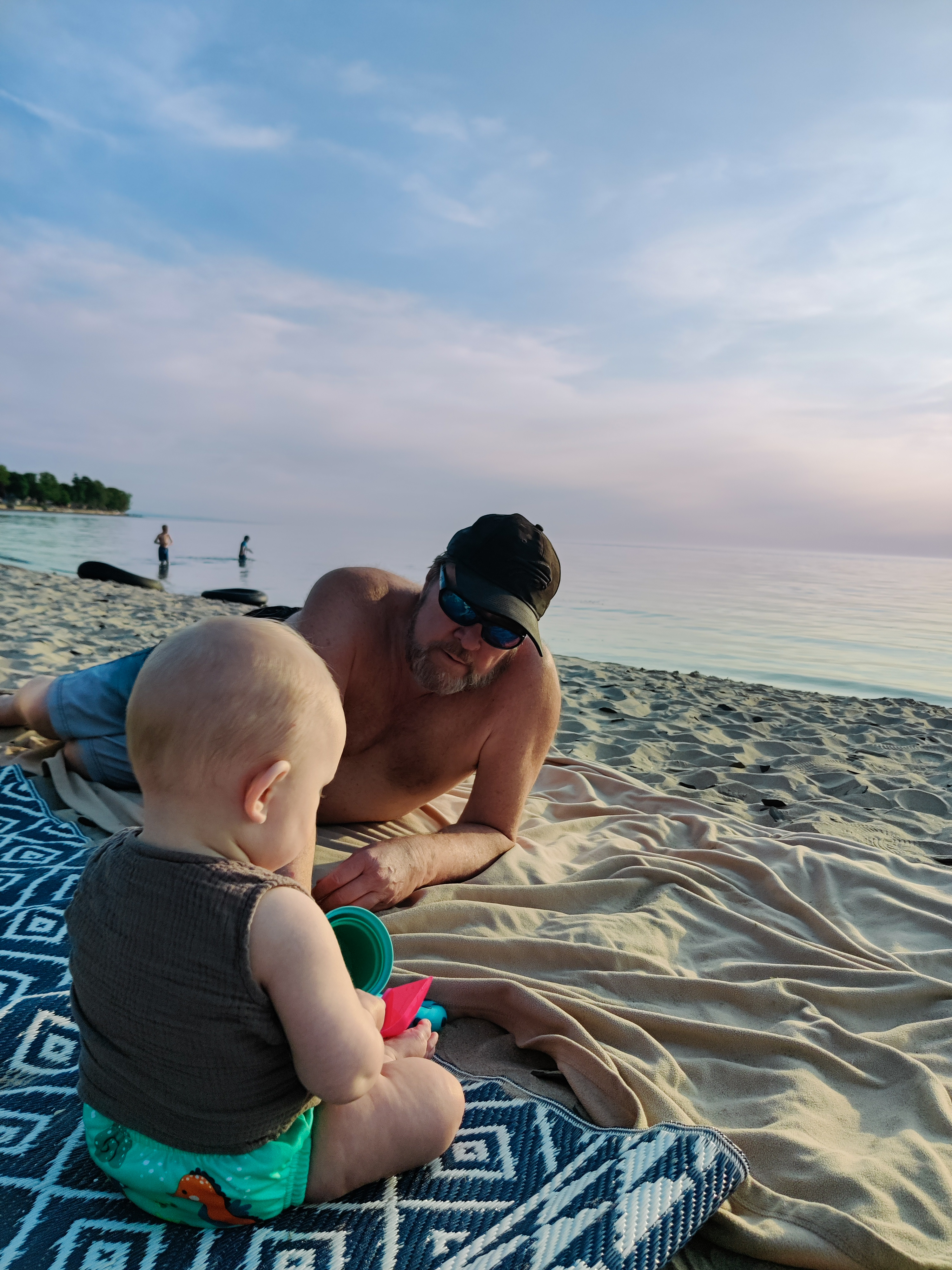 Larry shirtless on the beach at sunset, with baby Nova on a blanket, 2023