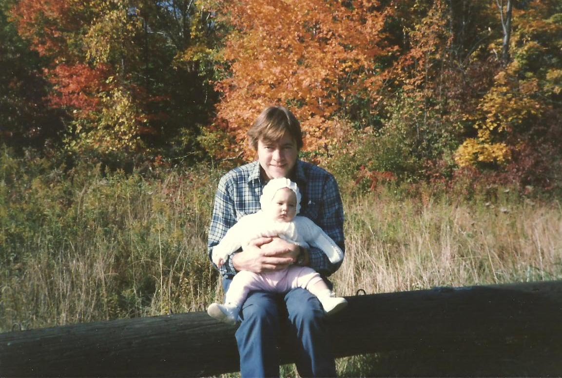 Larry holding baby Christina by a log fence in autumn, 1991