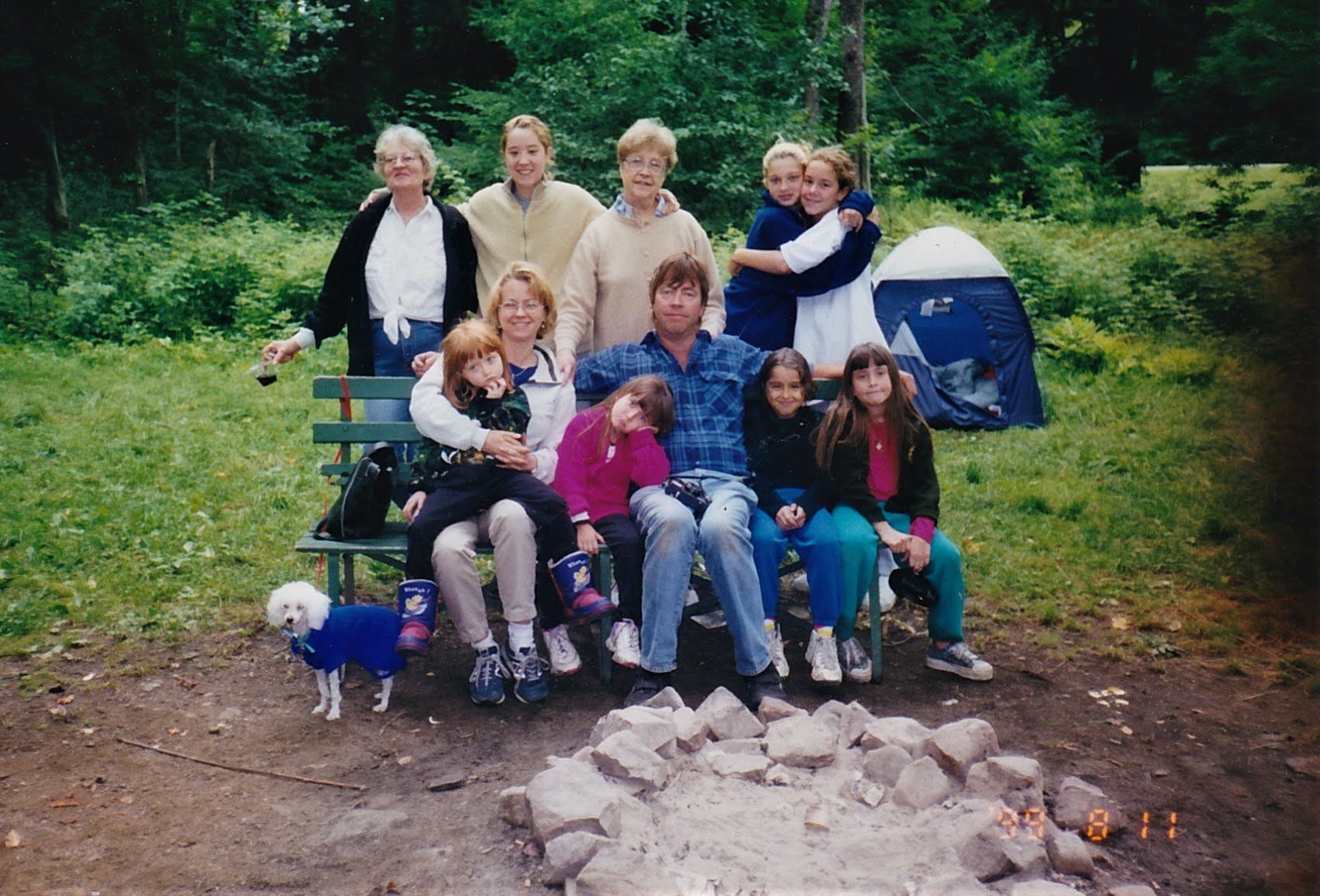 The family camping at Allegany State Park, August 1999, with a tent in the background, a fire pit in the foreground, and a dog in a blue jacket