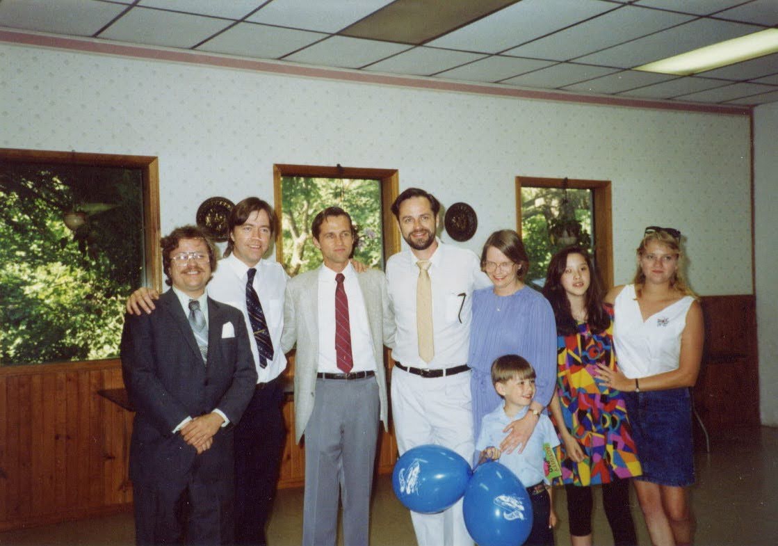 A Hilburger family gathering: eight adults in dressy clothes, a young boy in front holding two blue balloons